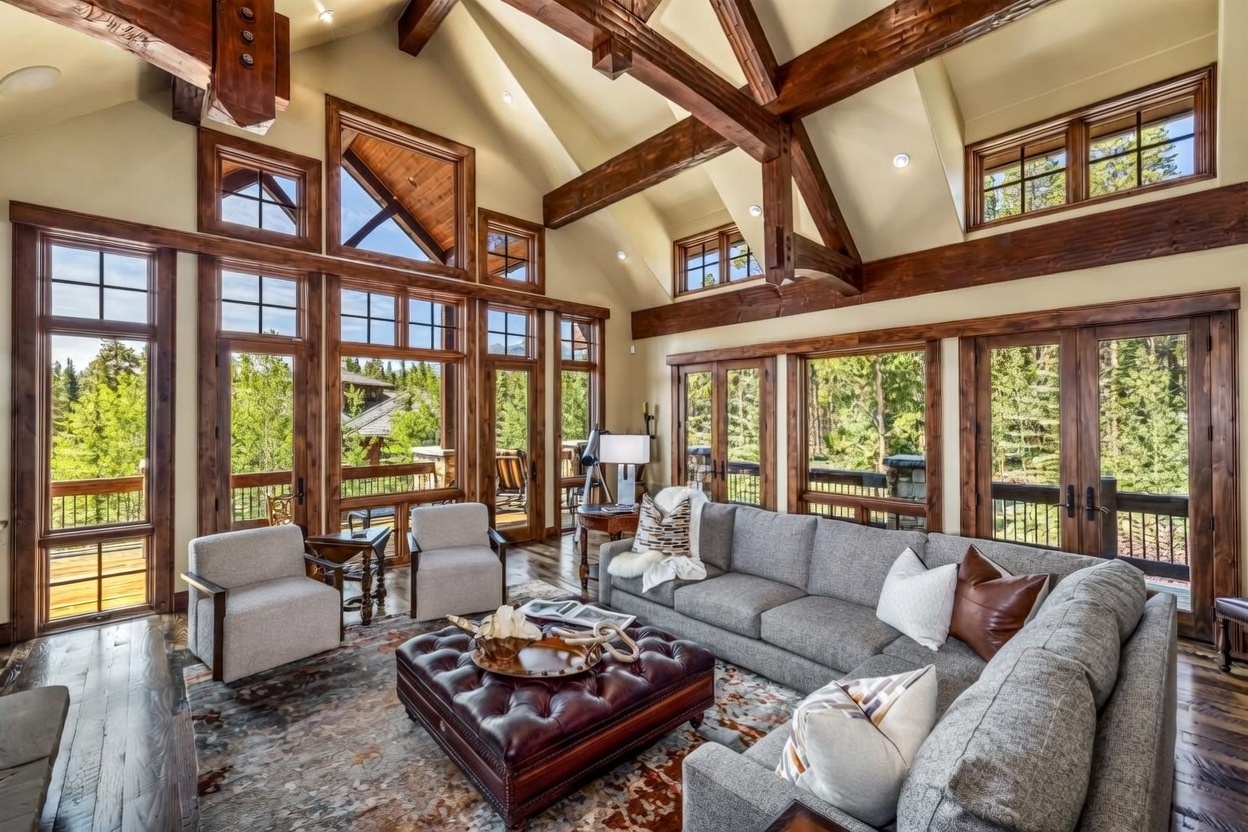This spacious living room features a vaulted ceiling with exposed timber beams and a large, dark leather tufted ottoman centered on a patterned area rug. A plush gray sectional sofa and two matching armchairs provide ample seating, while expansive windows offer panoramic views of the surrounding forest. The room is bathed in natural light, highlighting the warm wood trim and the sophisticated, rustic mountain aesthetic.