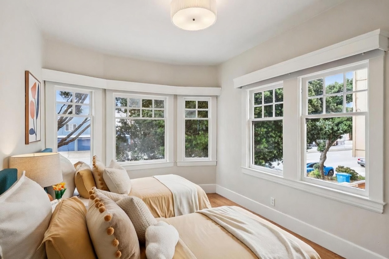 This bright and airy bedroom features two twin beds dressed in neutral-toned bedding with decorative pom-pom accents. A large bay window configuration and additional side windows flood the room with natural light, creating a welcoming and peaceful atmosphere. The space is finished with light-colored walls, hardwood flooring, and a simple, modern ceiling light fixture.