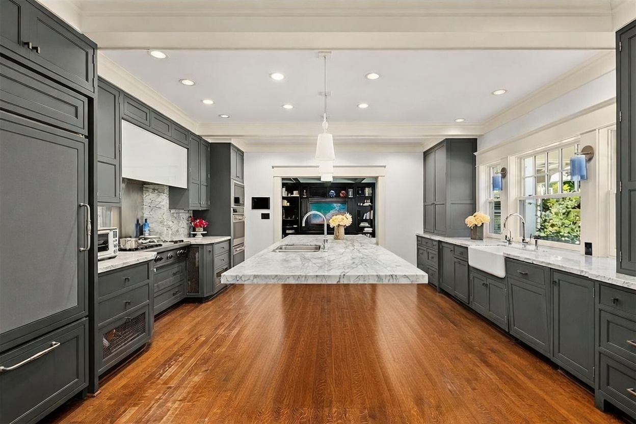 This spacious, high-end kitchen features dark-toned cabinetry contrasted by a large central island topped with a light-colored stone surface. The room is anchored by warm-toned wood flooring and illuminated by recessed lighting and a central pendant fixture, creating a sophisticated and inviting atmosphere. A view through the doorway reveals a secondary living space, emphasizing the open-concept layout of the home.