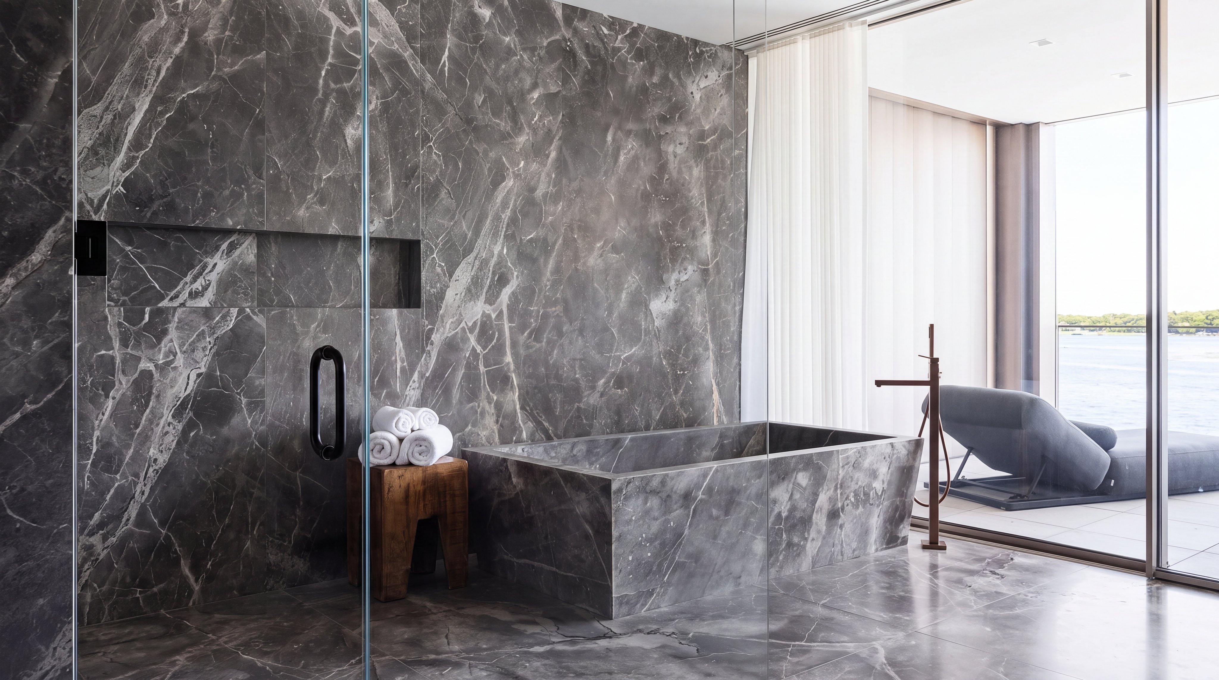 This luxurious bathroom features floor-to-ceiling dark grey marble paneling and a matching stone soaking tub. A glass partition separates the shower from the rest of the room, while a wooden stool holds folded white towels for a spa-like touch. The space opens up to a private balcony overlooking the ocean, emphasizing a high-end, serene retreat atmosphere.