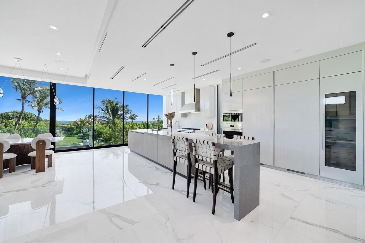 This sleek, modern kitchen features a large central island with a stone countertop and integrated seating, complemented by minimalist white cabinetry and high-end built-in appliances. The space is defined by expansive floor-to-ceiling windows that provide an unobstructed view of the lush, tropical greenery outside. The bright, open-concept design is enhanced by polished white flooring and contemporary pendant lighting, creating a sophisticated and airy atmosphere.