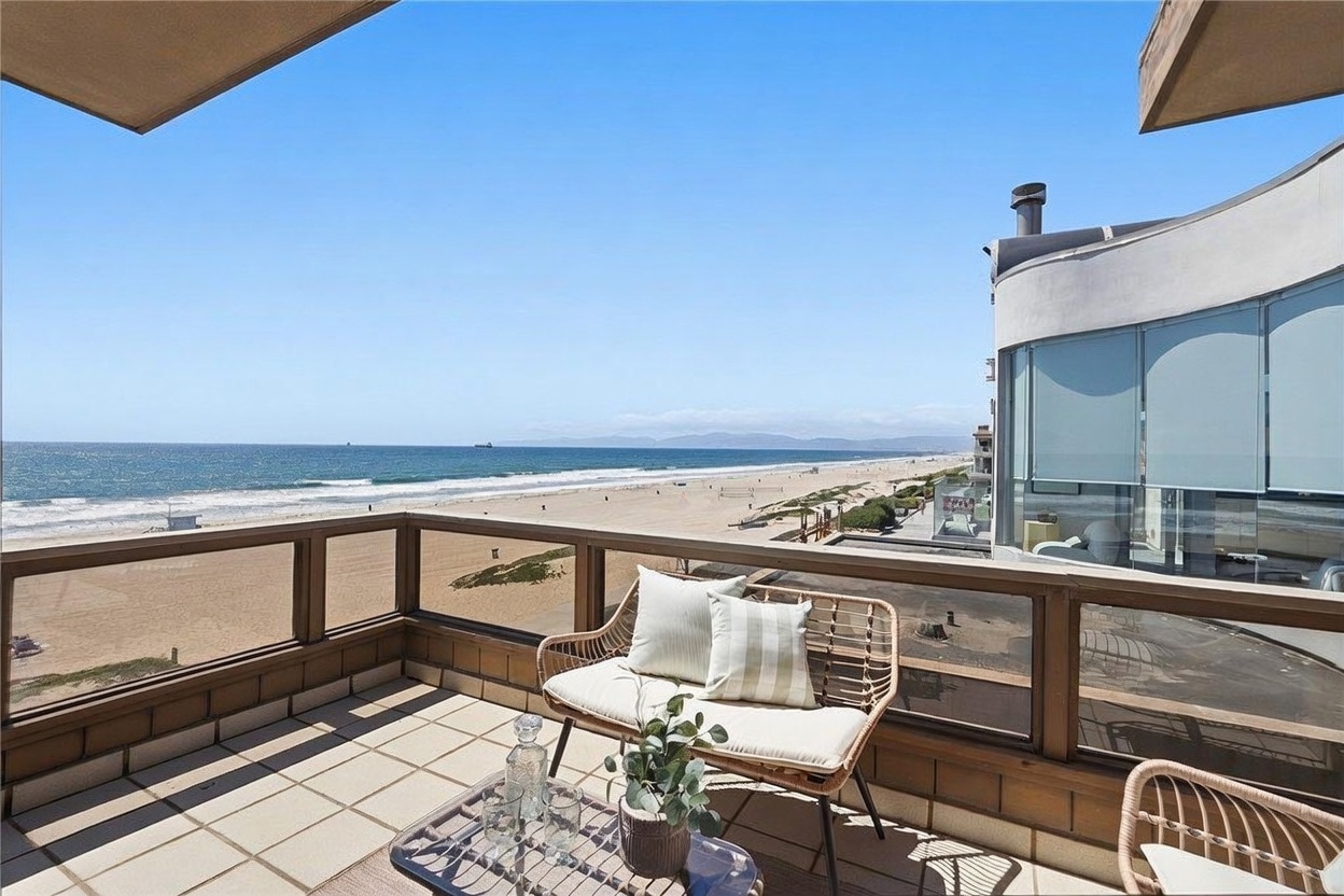 This elevated balcony offers a stunning, unobstructed panoramic view of the sandy beach and the Pacific Ocean. The space is furnished with a light-toned wicker loveseat and a small metal-framed table, creating a serene outdoor seating area. The balcony features a simple wood-framed railing with glass panels, allowing for an immersive connection with the coastal landscape.