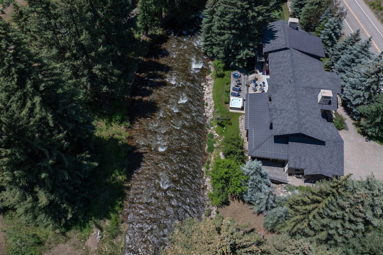 This high-angle aerial perspective captures a multi-story mountain home nestled beside a rushing river and dense forest. The property features a dark-toned roof, a spacious deck with outdoor seating, and a private hot tub overlooking the water. A paved road runs along the edge of the property, emphasizing its secluded yet accessible location in a natural, wooded setting.
