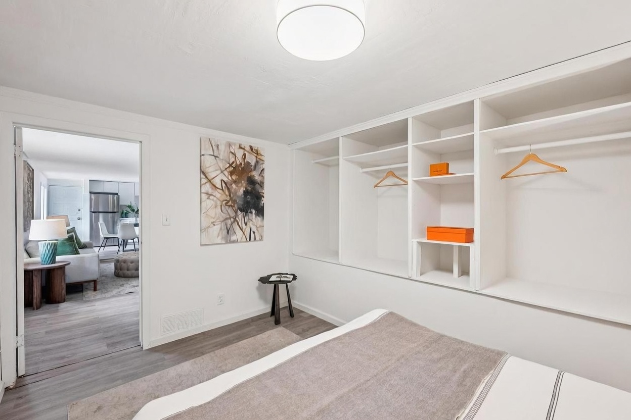 This bedroom features a minimalist design with white walls, light wood-look flooring, and a built-in open shelving unit that provides ample storage. A neutral-toned bed is positioned in the foreground, while an abstract piece of wall art adds a focal point near the doorway leading to an adjacent living area. The perspective captures the clean, bright aesthetic of the room, highlighting the seamless transition between living spaces.