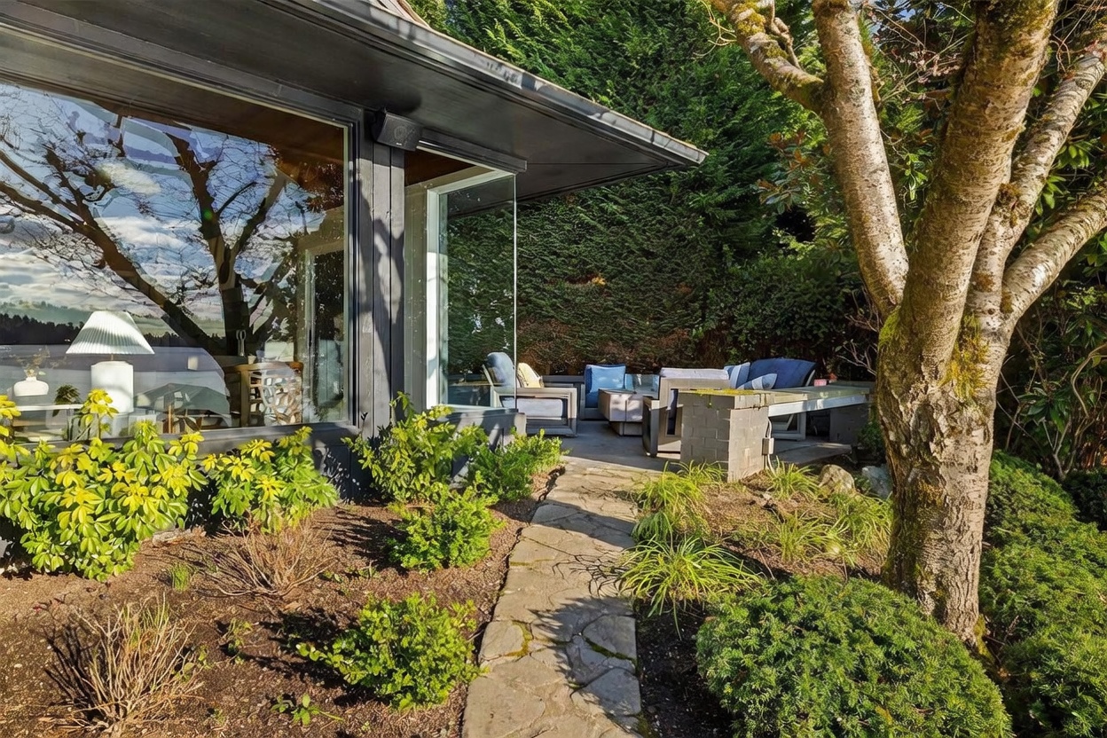 This image captures a serene outdoor patio area adjacent to a modern home with expansive floor-to-ceiling glass windows. The space features comfortable outdoor seating, a stone pathway leading through lush landscaping, and a mature tree providing natural shade. The perspective highlights the seamless transition between the indoor living space and the private, tranquil backyard retreat.