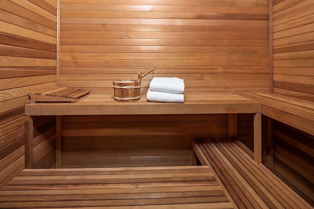 This image showcases a traditional sauna interior constructed entirely from light-toned wood paneling. The space features tiered wooden benches for seating, a wooden bucket with a ladle, and a neatly folded white towel, creating a serene and relaxing atmosphere. The perspective is a straight-on, eye-level shot that highlights the warm, natural textures and the compact, functional design of the wellness space.
