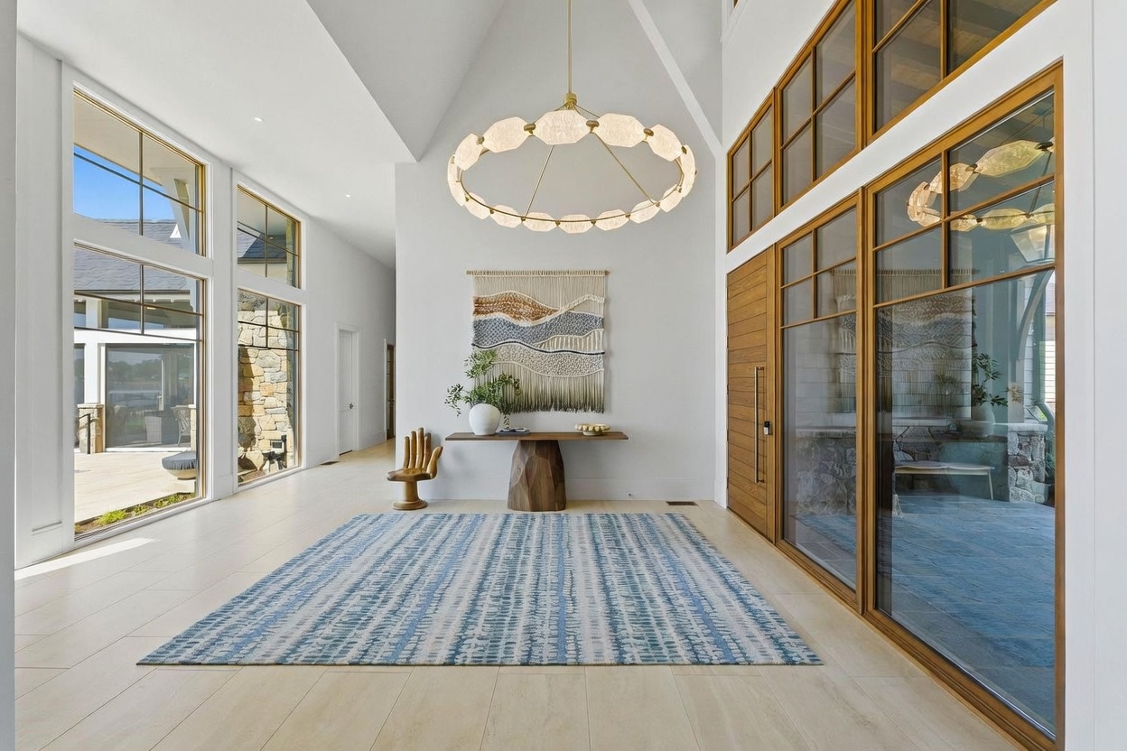 This grand, light-filled entryway features soaring vaulted ceilings and expansive glass walls that provide a seamless connection to the outdoors. The space is anchored by a large, textured blue and white area rug, a sculptural wooden console table, and a unique circular chandelier with translucent, organic-shaped pendants. The minimalist aesthetic is complemented by a woven wall hanging and a distinctive hand-shaped wooden chair, creating a sophisticated and welcoming atmosphere.