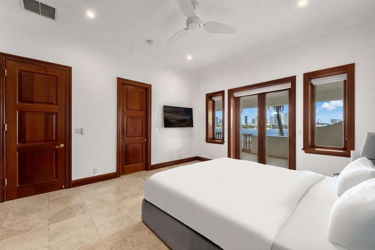 This bright and airy primary bedroom features neutral walls, light-colored tile flooring, and a ceiling fan. A large glass door and adjacent windows provide access to a balcony with a scenic water view, filling the room with natural light. The space is furnished with a bed and a wall-mounted television, creating a clean and tranquil atmosphere.