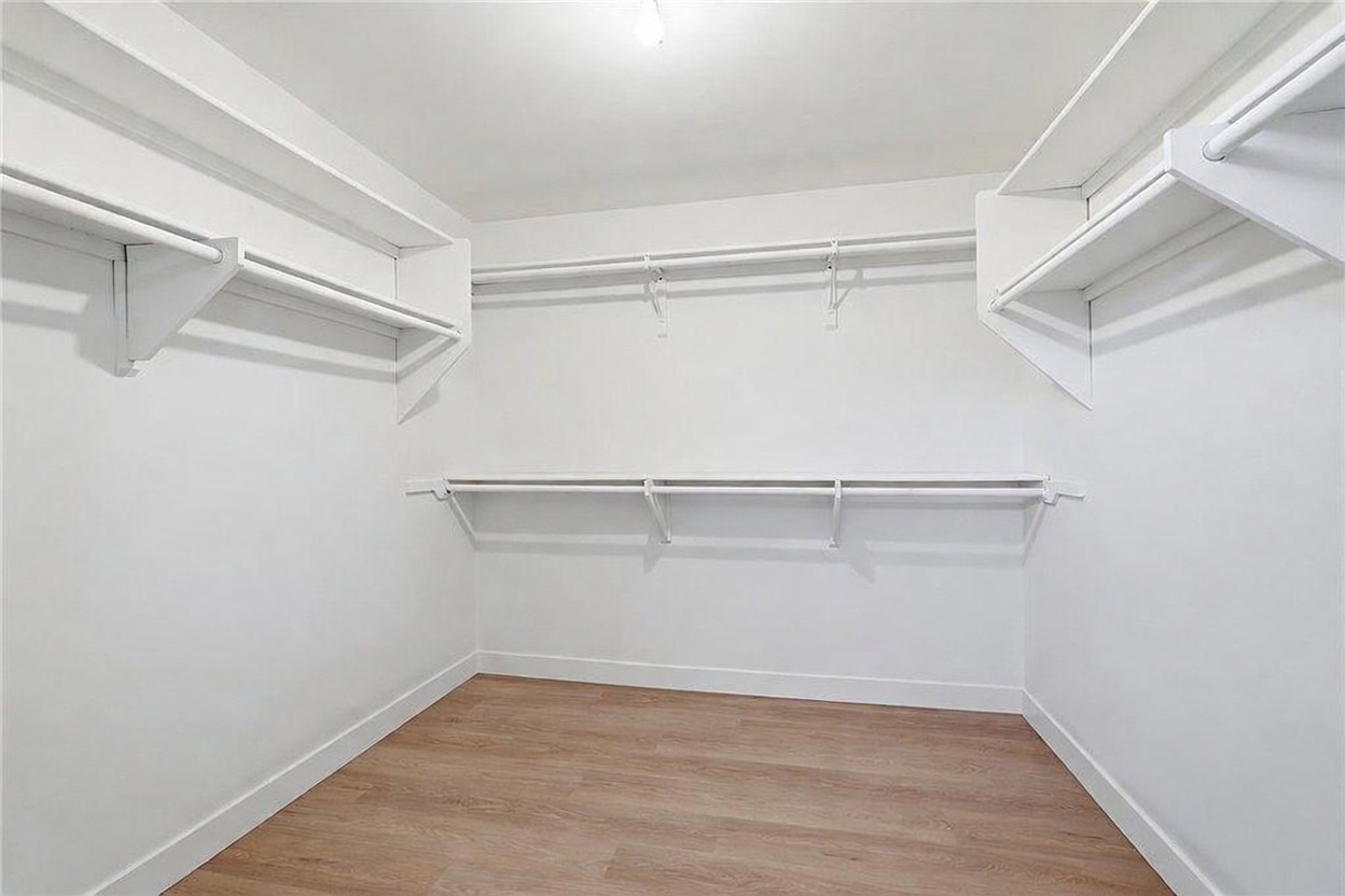 This walk-in closet features a clean, minimalist design with white walls and light wood-toned flooring. The space is equipped with multiple levels of white shelving and hanging rods, providing ample storage and organization options. The perspective is centered, showcasing the depth and functional layout of the closet.