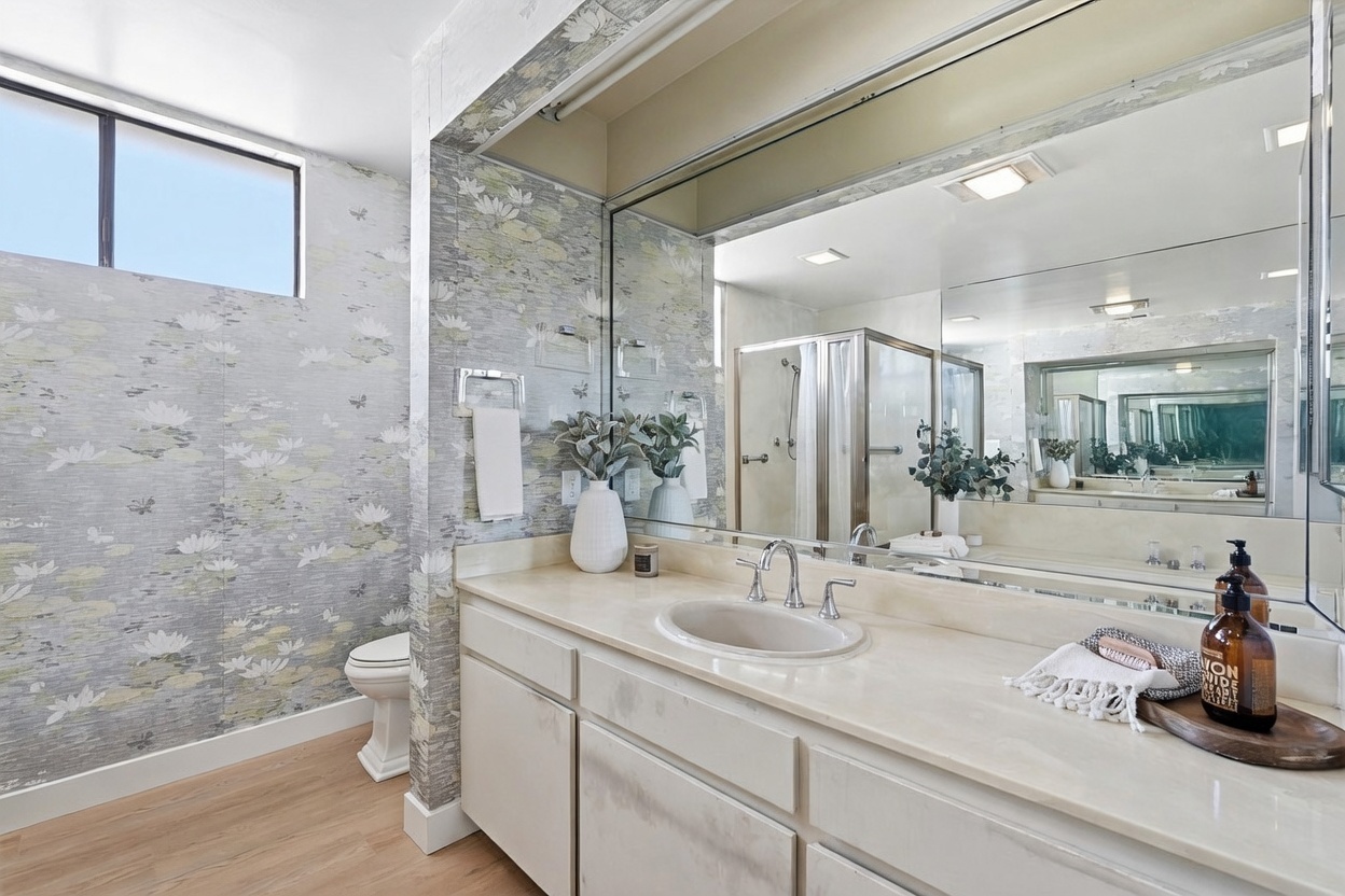 This bathroom features a vanity with a light-toned countertop and a large, wide mirror that reflects the shower area. The walls are adorned with a botanical-themed wallpaper, and the space includes a toilet positioned near a high-placed window. The overall aesthetic is bright and functional, with light wood-look flooring completing the clean, neutral design.