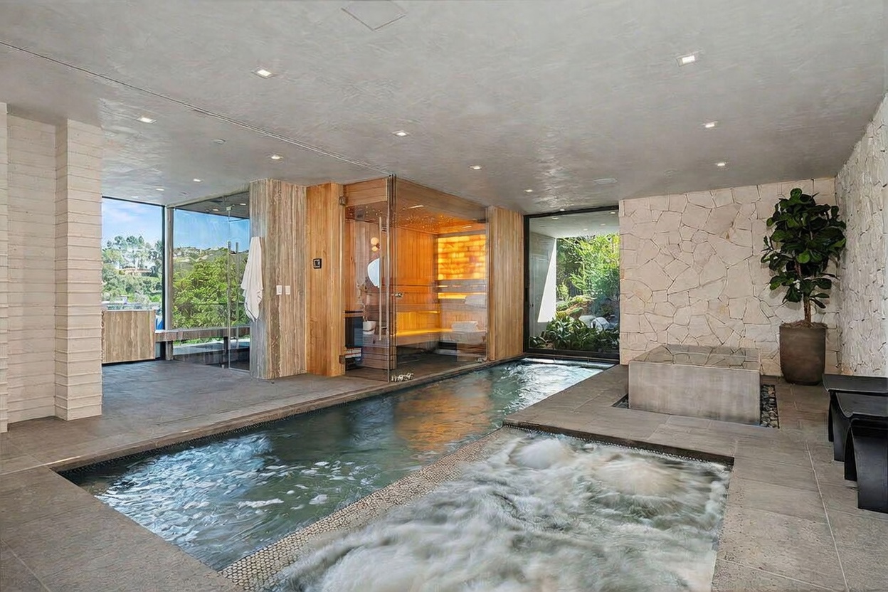 This luxurious indoor spa area features a long lap pool adjacent to a bubbling hot tub, set against a backdrop of natural stone walls and light wood paneling. A glass-enclosed sauna with warm interior lighting serves as a focal point, while large windows provide a view of the lush greenery outside. The space is designed for relaxation, combining high-end materials and modern architectural elements to create a serene, resort-like atmosphere.