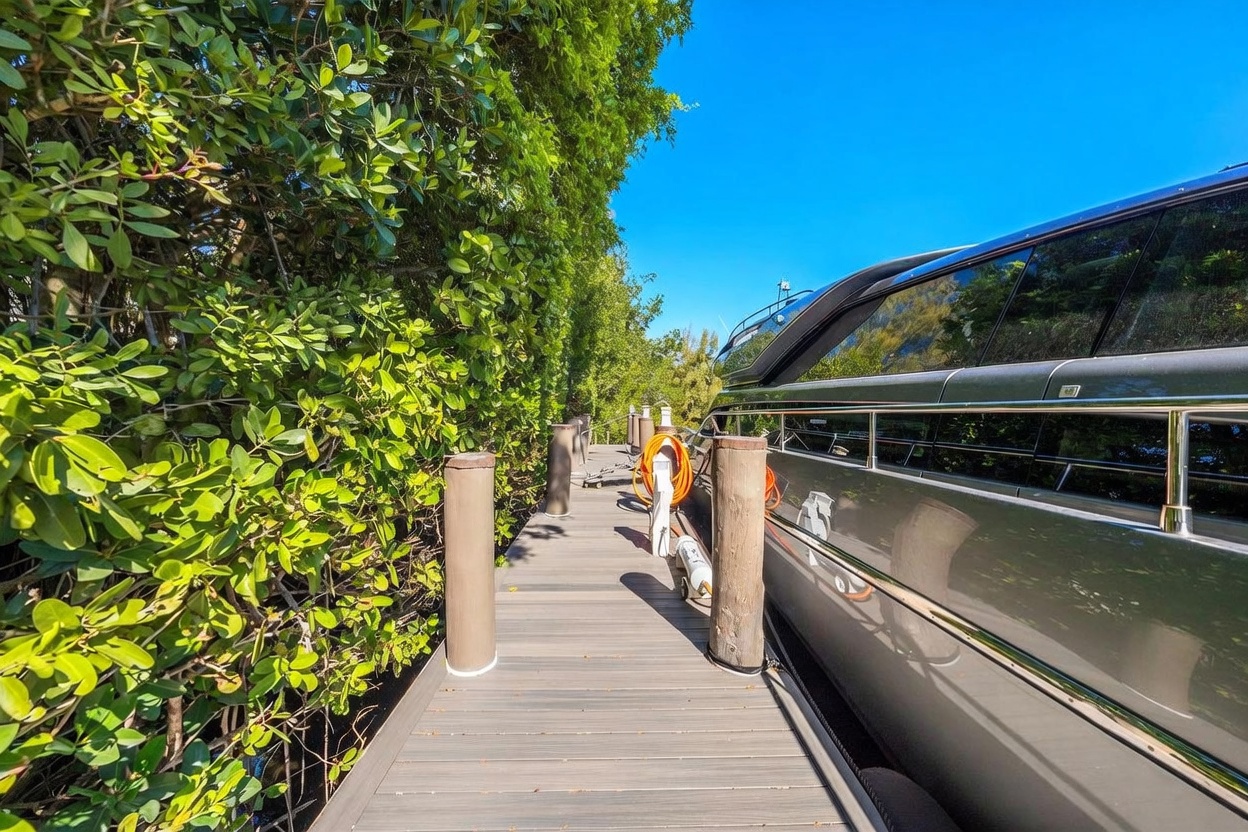 This image captures a private wooden dock extending alongside a large, sleek yacht moored in a tranquil, lush setting. The perspective is from the dock looking forward, showcasing the contrast between the natural, dense green foliage on the left and the polished, dark metallic hull of the boat on the right. The scene conveys a sense of luxury and peaceful waterfront living, with a bright blue sky overhead.