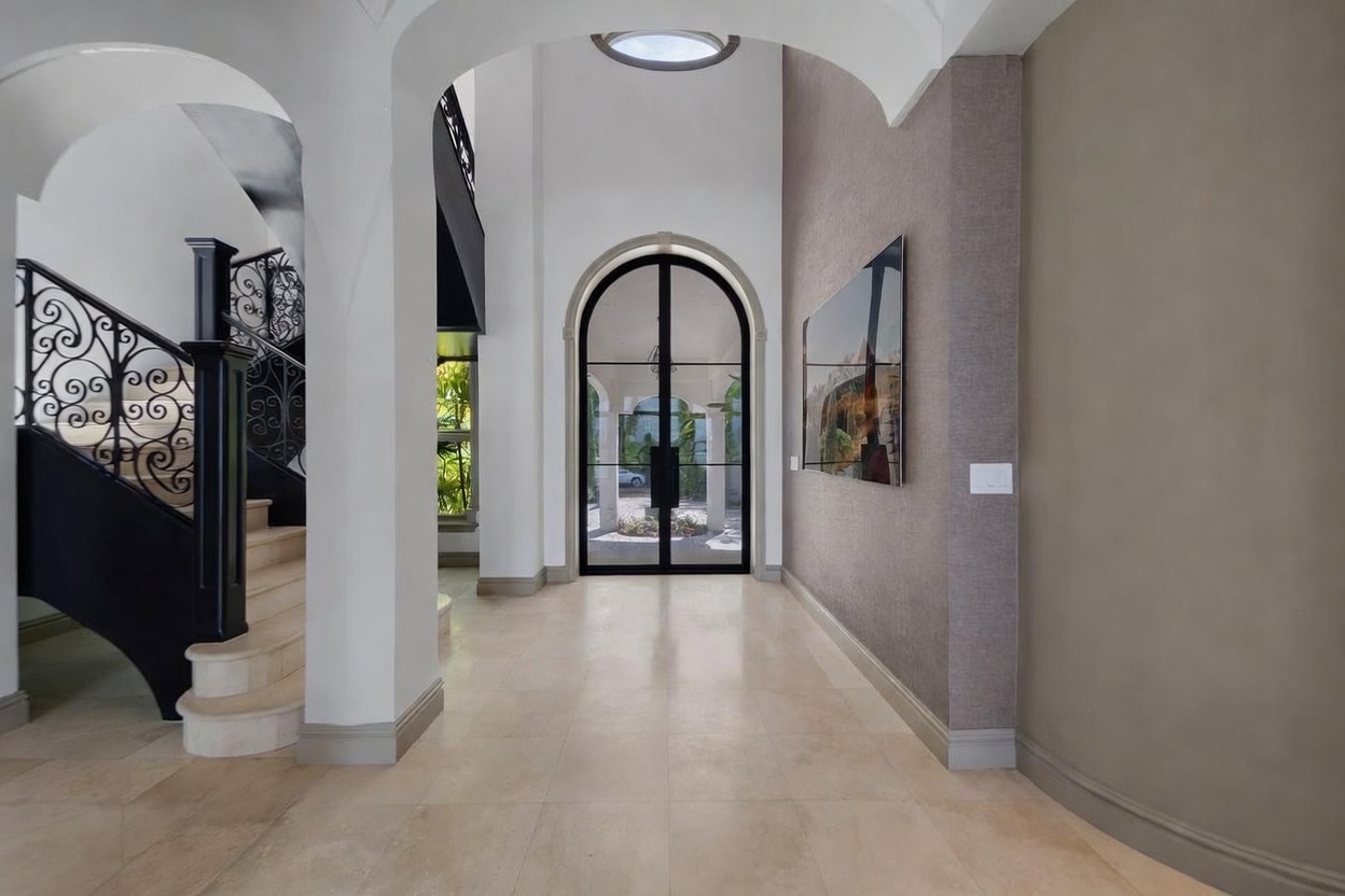 This elegant foyer features a grand staircase with intricate black wrought-iron railings and a polished wood handrail. The space is anchored by large, light-toned stone floor tiles and leads toward a striking arched glass door that provides a view of the outdoor courtyard. A textured, neutral-toned accent wall displays a large piece of art, creating a sophisticated and welcoming transition area.
