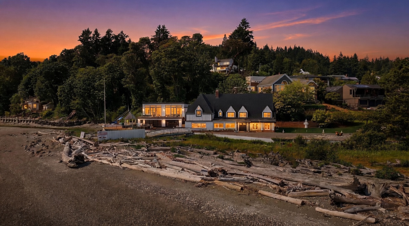 This stunning waterfront property is captured at dusk, showcasing a unique architectural blend of a traditional gabled home and a modern, glass-walled addition. The house sits directly on a beach scattered with driftwood, with a private bridge leading to the entrance. The vibrant orange and purple sunset provides a dramatic backdrop to the lush, tree-lined hillside and the illuminated windows of the residence.