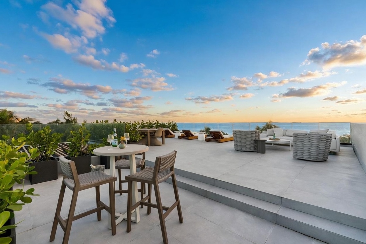 This expansive rooftop terrace offers a luxurious outdoor living space with panoramic ocean views under a clear blue sky. The area is furnished with a high-top dining set in the foreground and a comfortable lounge seating arrangement with plush sofas and woven chairs further back. A glass railing provides an unobstructed view of the horizon, creating a seamless connection between the terrace and the sea.
