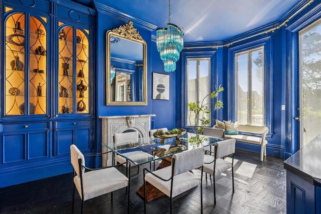 This vibrant dining room is defined by its bold, monochromatic blue walls and matching built-in cabinetry with glass-fronted display shelves. A glass-topped dining table sits at the center, surrounded by modern, light-colored upholstered chairs, while a striking blue tiered chandelier hangs above. The room features a classic stone fireplace mantel, a large ornate gold-framed mirror, and dark wood flooring, creating a sophisticated and dramatic atmosphere.