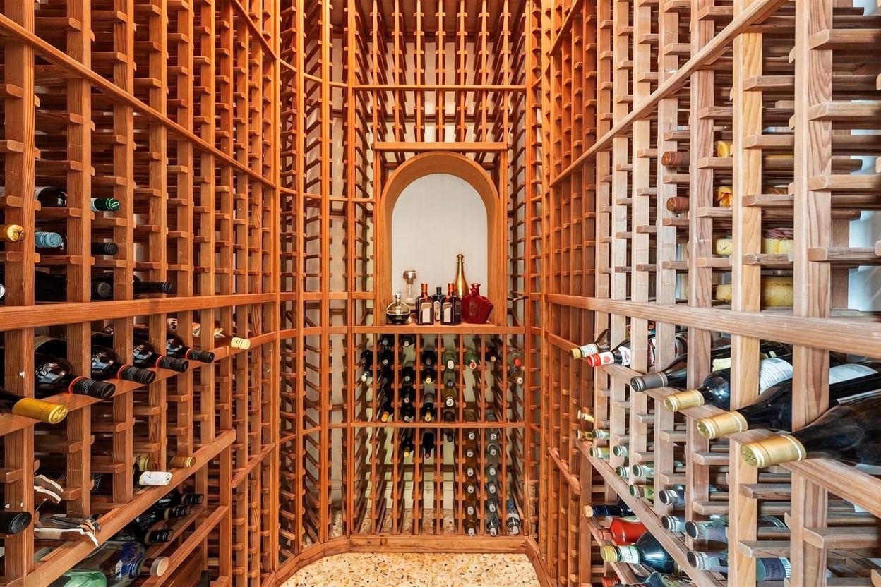 This dedicated wine cellar features extensive custom wood racking that spans the walls, providing ample storage for a large collection of bottles. A central arched alcove serves as a focal point, displaying select bottles and decorative items on a shelf. The space is finished with a light-colored, speckled stone floor, creating a sophisticated and organized environment for wine storage.