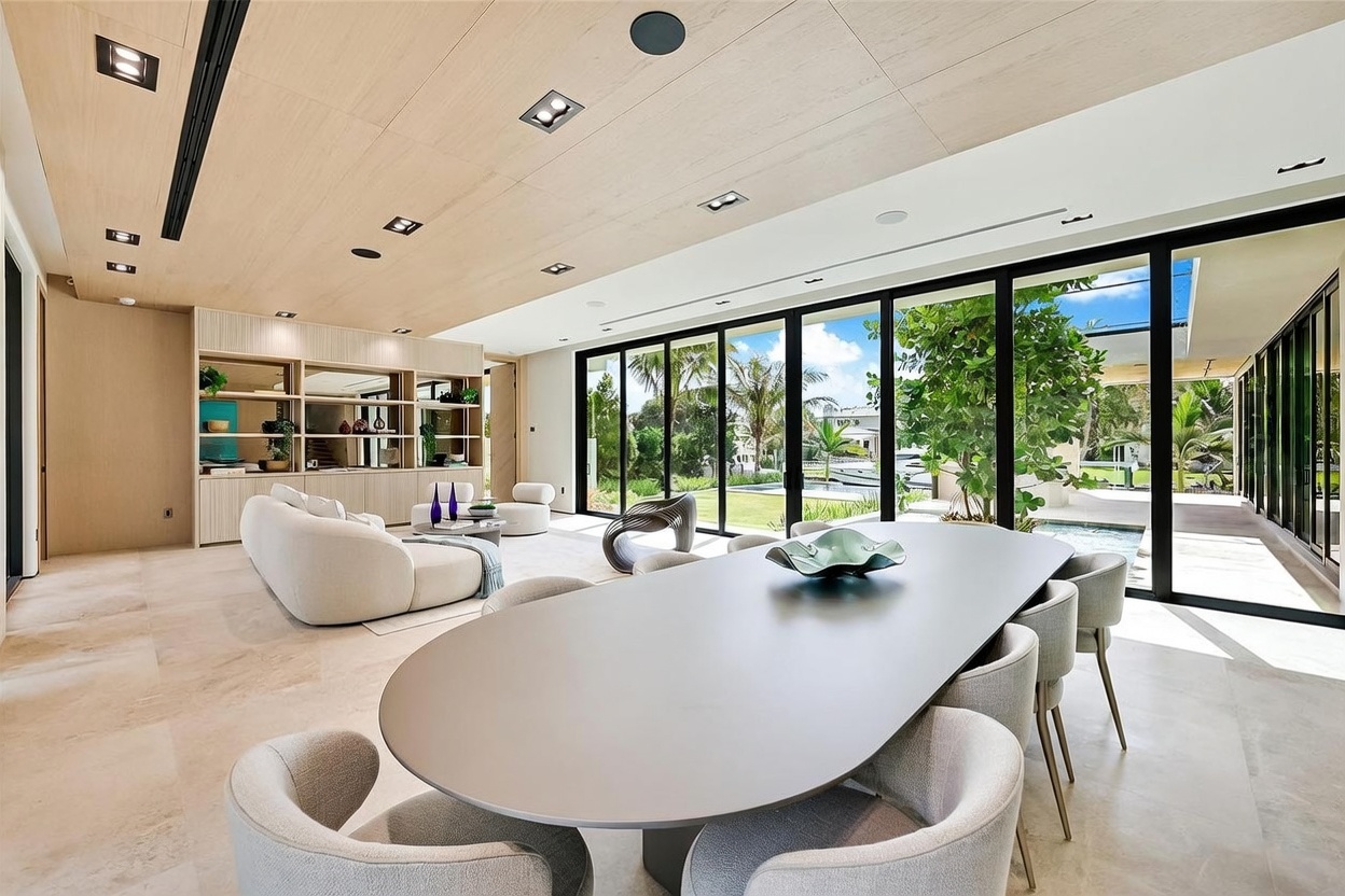 This expansive, open-concept living and dining area features a modern aesthetic with light wood-paneled ceilings and large, floor-to-ceiling glass walls that provide seamless indoor-outdoor flow. The dining space is anchored by a sleek, oval-shaped table surrounded by upholstered chairs, while the adjacent living area includes a curved sofa and custom built-in shelving. The room is bathed in natural light, offering a clear view of the lush backyard, swimming pool, and waterfront beyond.