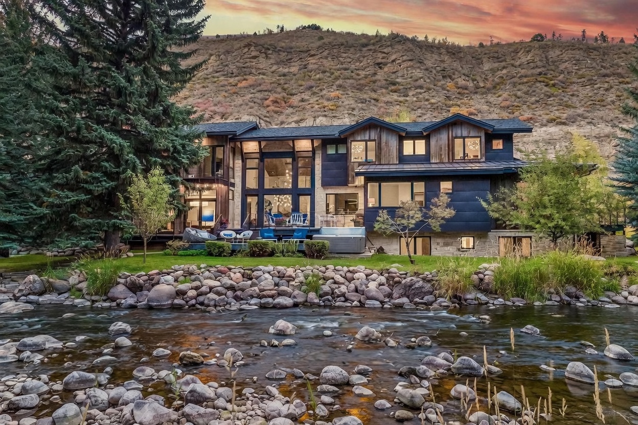 This stunning mountain residence features a contemporary architectural design with a mix of dark metal siding and warm wood accents. The home is positioned along a rocky riverbank, with a spacious deck area that includes outdoor seating and a hot tub. Large windows dominate the rear facade, reflecting the dramatic sunset sky and the rugged hillside backdrop.
