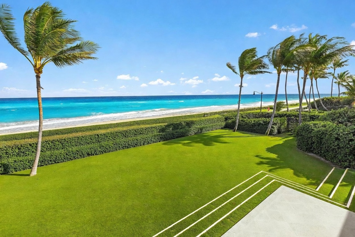 This elevated view captures a pristine, manicured lawn leading directly to a private beach and the turquoise ocean beyond. Tall palm trees frame the scene, adding a tropical aesthetic to the expansive outdoor space. The foreground features a clean, paved patio area with geometric lines, emphasizing a seamless transition between the residence and the coastal landscape.