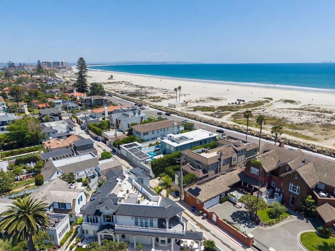 This high-angle aerial view captures a coastal neighborhood situated directly adjacent to a wide, sandy beach and the Pacific Ocean. The scene showcases a mix of residential architectural styles, including multi-story homes with flat and pitched roofs, some featuring rooftop decks and private pools. The perspective emphasizes the prime location, highlighting the proximity to the shoreline and the surrounding suburban landscape under a clear blue sky.