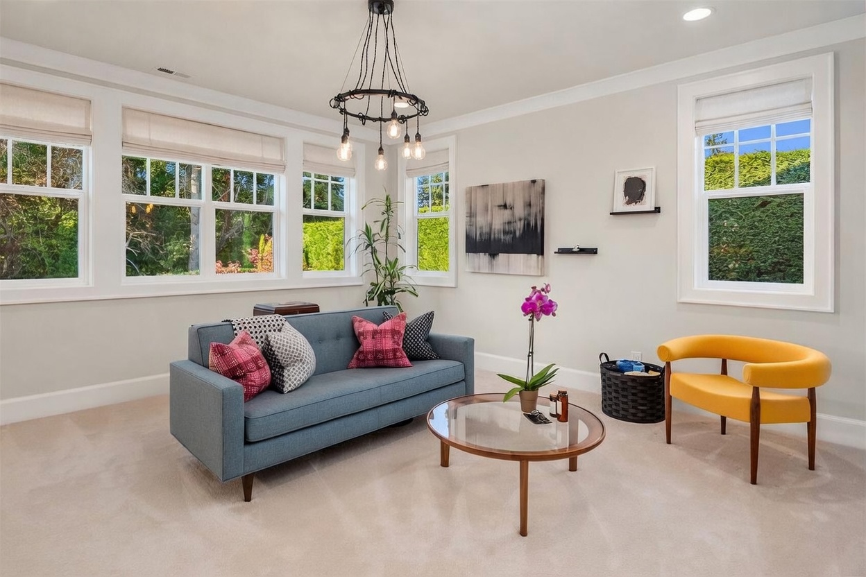 This bright and inviting living room features a mid-century modern aesthetic with a blue sofa accented by patterned pillows and a vibrant yellow armchair. A circular glass-topped coffee table sits in the center, complemented by a unique multi-bulb chandelier hanging from the ceiling. Large windows line the walls, allowing ample natural light to fill the space and providing a pleasant view of the surrounding greenery.