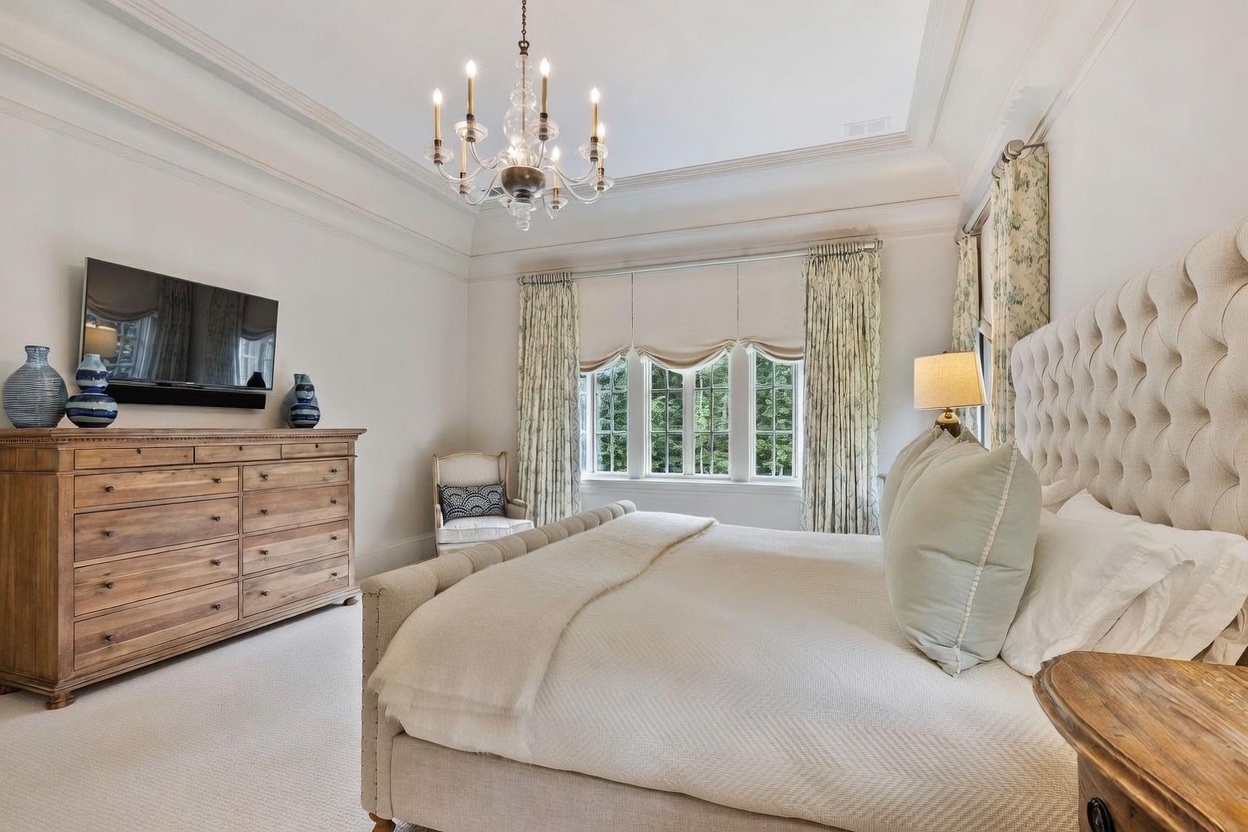 This elegant primary bedroom features a large, tufted upholstered bed as the focal point, complemented by a light wood dresser and a classic crystal chandelier hanging from a tray ceiling. Tall windows dressed in patterned curtains allow natural light to fill the space, creating a serene and sophisticated atmosphere. The room is finished with neutral carpeting and a small accent chair, offering a comfortable and refined retreat.