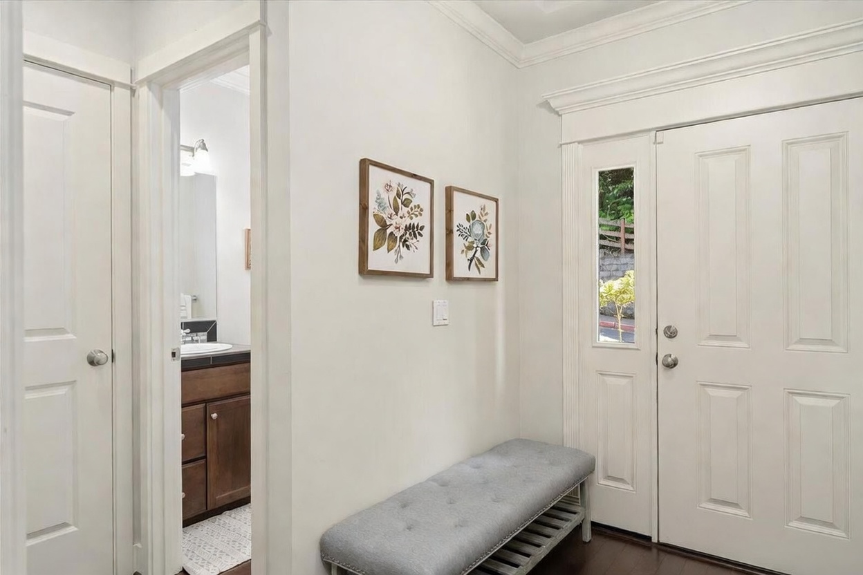 This bright and welcoming entryway features a white front door with a vertical glass panel and a matching gray upholstered bench placed against the wall. Two framed floral botanical prints hang above the bench, adding a touch of charm to the neutral space. A doorway to the left provides a glimpse into a bathroom with dark cabinetry and a light-colored countertop, creating a clean and cohesive transition between rooms.