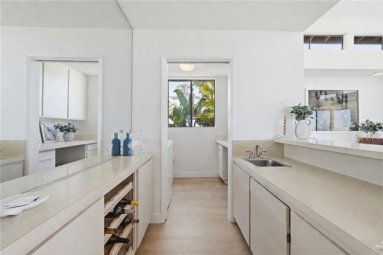 This bright, contemporary kitchen area features a wet bar with a built-in wine rack and a sink set into a long, light-toned countertop. The space is characterized by clean white cabinetry and light wood-look flooring, creating an airy and cohesive aesthetic. A doorway leads into an adjacent laundry room, while a pass-through window provides a view into another living space, enhancing the open-concept feel.