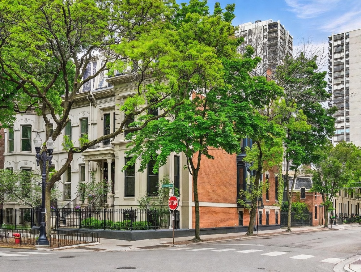 This image captures a historic, multi-story brick townhouse situated on a corner lot, showcasing classic architectural details and ornate masonry. The property is framed by mature, lush green trees and features a decorative black iron fence enclosing the front yard. The perspective is a street-level view, highlighting the building's prominent position at a quiet intersection with a stop sign.
