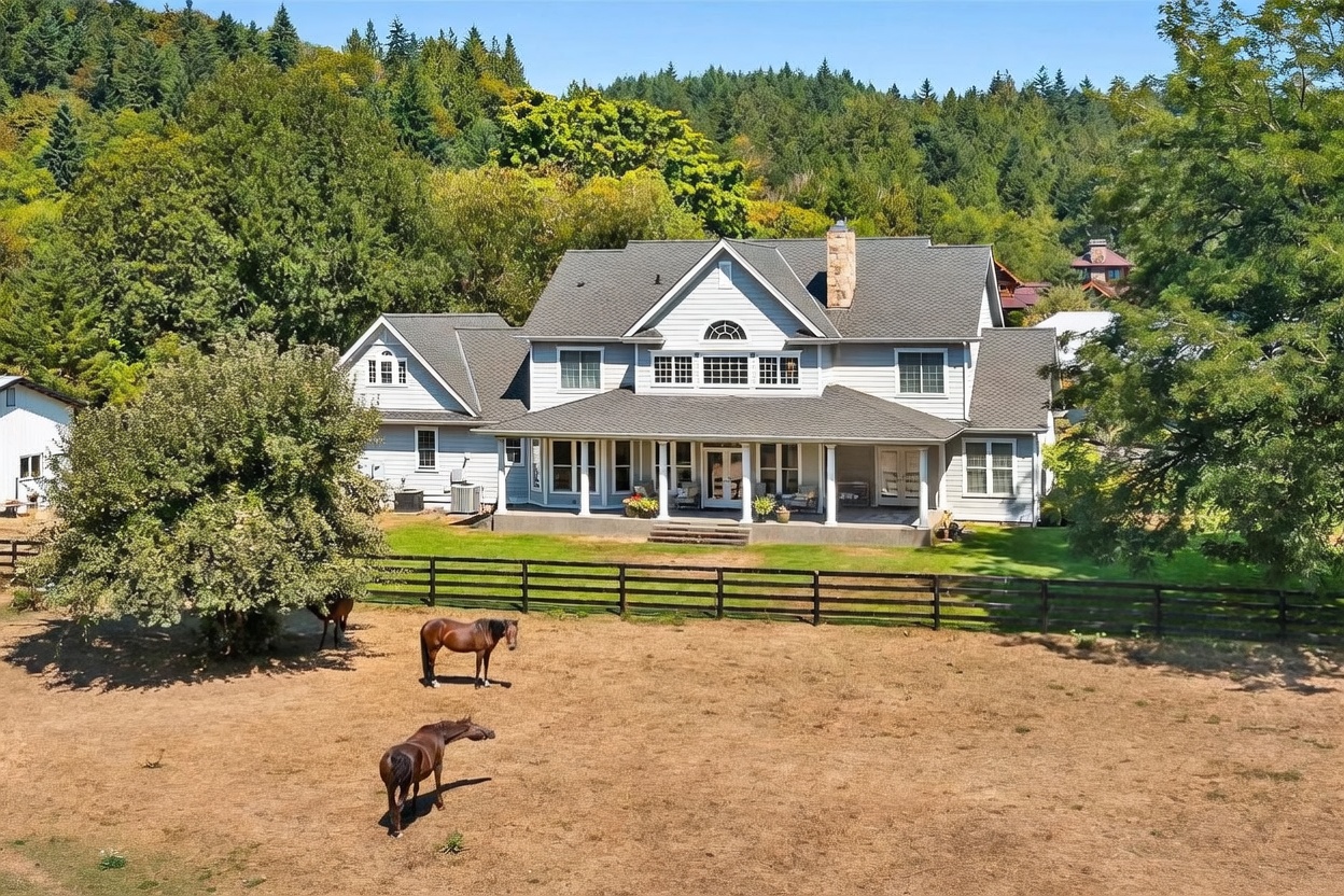 This expansive rear view of a large, multi-story suburban home showcases a classic architectural style with light-colored siding and a prominent stone chimney. A wide, covered porch spans the back of the house, providing a shaded transition to the grassy lawn, which is bordered by a dark wooden fence. In the foreground, several horses graze in a dry, open pasture, emphasizing the rural or equestrian nature of the property set against a backdrop of dense, mature forest.