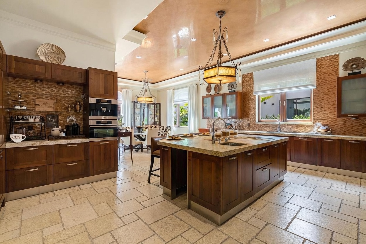 This spacious, high-end kitchen features rich, dark wood cabinetry, a large central island with a stone countertop, and a striking coffered ceiling with a glossy finish. The room is illuminated by elegant pendant lighting and natural light streaming through large windows, creating a warm and inviting atmosphere. The perspective is a wide-angle shot from the kitchen floor, showcasing the seamless flow between the cooking area and the adjacent dining space.