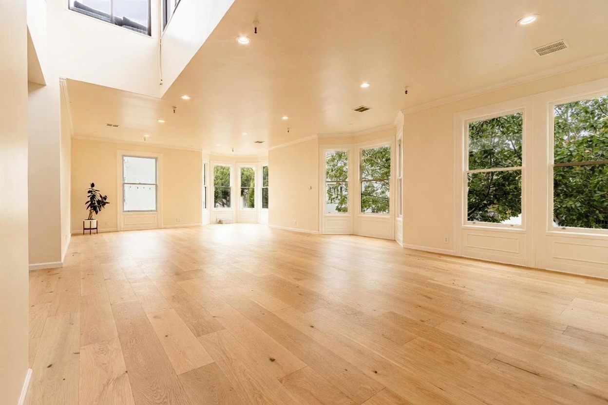 This spacious, open-concept living area features light wood flooring and a neutral color palette, creating a bright and airy atmosphere. The room is defined by multiple large windows, including a bay window configuration that invites abundant natural light into the space. A high, vaulted ceiling adds to the sense of volume, while recessed lighting provides a clean and modern aesthetic.