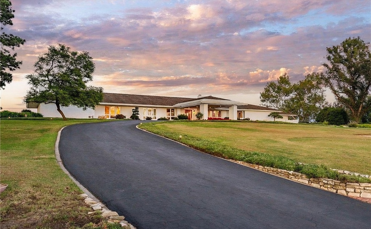 This wide-angle shot captures the front exterior of a sprawling, single-story ranch-style home set against a dramatic, colorful sunset sky. A long, curved asphalt driveway leads up to the main entrance, which is sheltered by a prominent covered portico. The property features expansive manicured lawns, mature trees, and a low stone retaining wall along the driveway, creating a welcoming and established curb appeal.