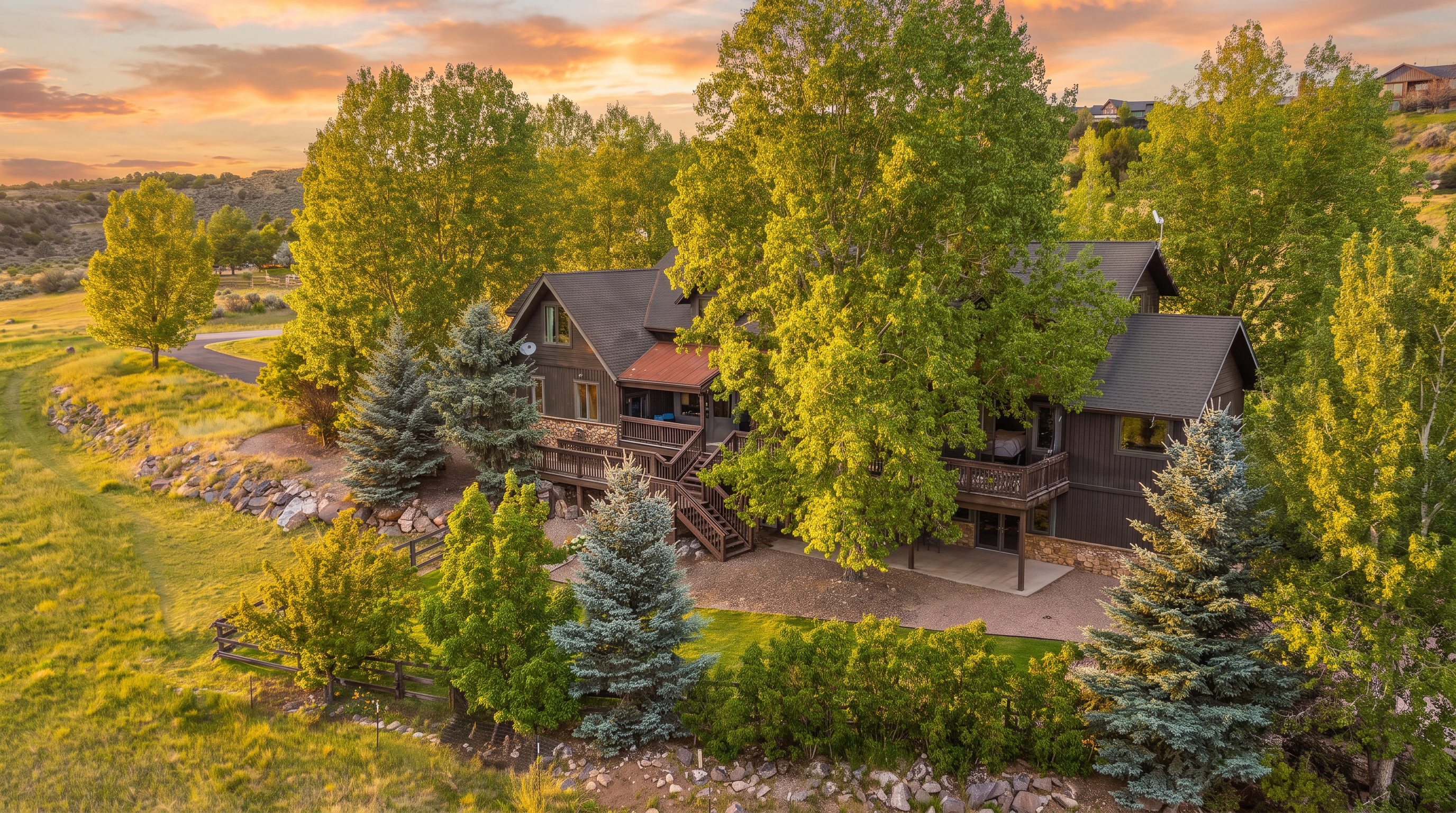 This high-angle drone shot captures a charming, rustic multi-story home nestled in a serene, semi-arid landscape characterized by expansive greenery and rocky terrain. The house features weathered wooden siding, a prominent metal-accented roof section, and extensive tiered wooden decking that seamlessly integrates with the natural surroundings. A manicured lawn and various evergreen trees provide a soft contrast to the rugged, dry hillside backdrop, contributing to an appealing mountain-retreat aesthetic.