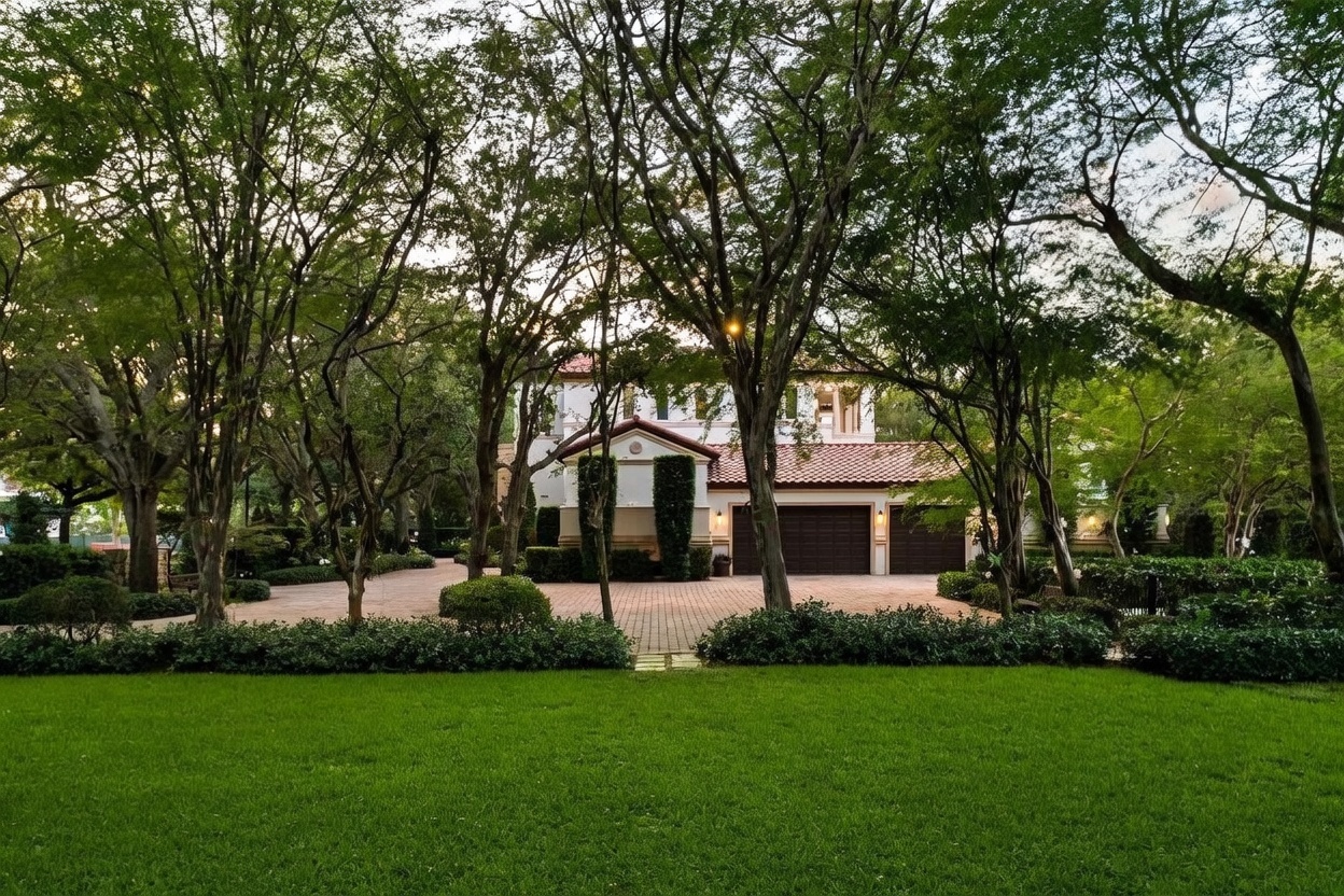 This front-facing view captures a stately Mediterranean-style residence nestled behind a lush canopy of mature trees. The home features a multi-car garage, a terracotta-tiled roof, and a paved driveway that leads to the main entrance. A well-manicured lawn in the foreground provides a sense of privacy and curb appeal, creating a serene and established atmosphere.