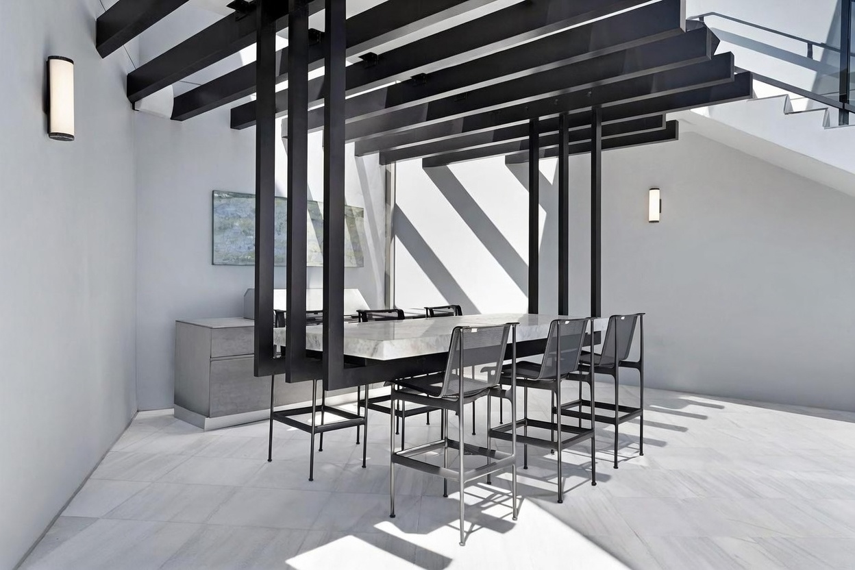 This modern outdoor dining area features a stone-topped table positioned beneath a striking black architectural pergola structure. The space is finished with light-toned stone flooring and minimalist metal bar stools, creating a clean, contemporary aesthetic. The perspective captures the geometric interplay of the overhead beams and the adjacent staircase, emphasizing a sophisticated, open-air design.