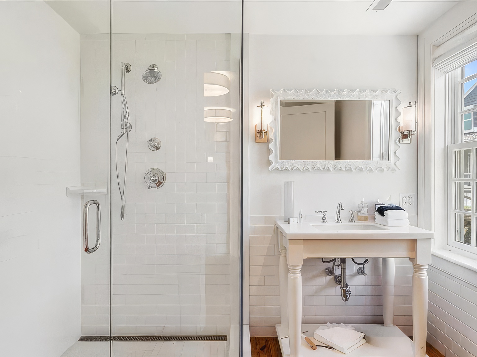 This bright, clean bathroom features a glass-enclosed walk-in shower with white subway tile walls and a modern shower system. To the right, a pedestal-style vanity with a white countertop and polished metal fixtures sits beneath a decorative framed mirror and wall-mounted sconces. The space is finished with white subway tile wainscoting and a window that allows natural light to fill the room, creating an airy and classic aesthetic.