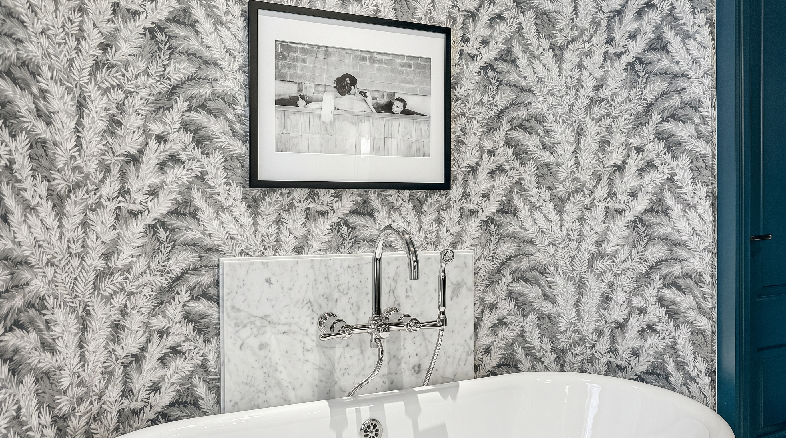 This elegant bathroom features a freestanding white soaking tub set against a striking wall covered in a detailed, monochromatic botanical-patterned wallpaper. A polished metal faucet is mounted on a rectangular stone panel behind the tub, while the floor is finished in large-format stone tiles. The space is framed by deep blue trim and doors, creating a sophisticated and high-contrast aesthetic.