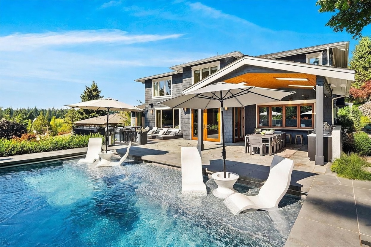 This luxurious outdoor living space features a modern swimming pool with a shallow tanning ledge equipped with white lounge chairs and umbrellas. A covered patio area provides a shaded dining space, while the dark-toned house exterior with large windows creates a sophisticated, contemporary aesthetic. The scene is captured from a low angle, emphasizing the seamless transition between the pool deck and the home's architecture.