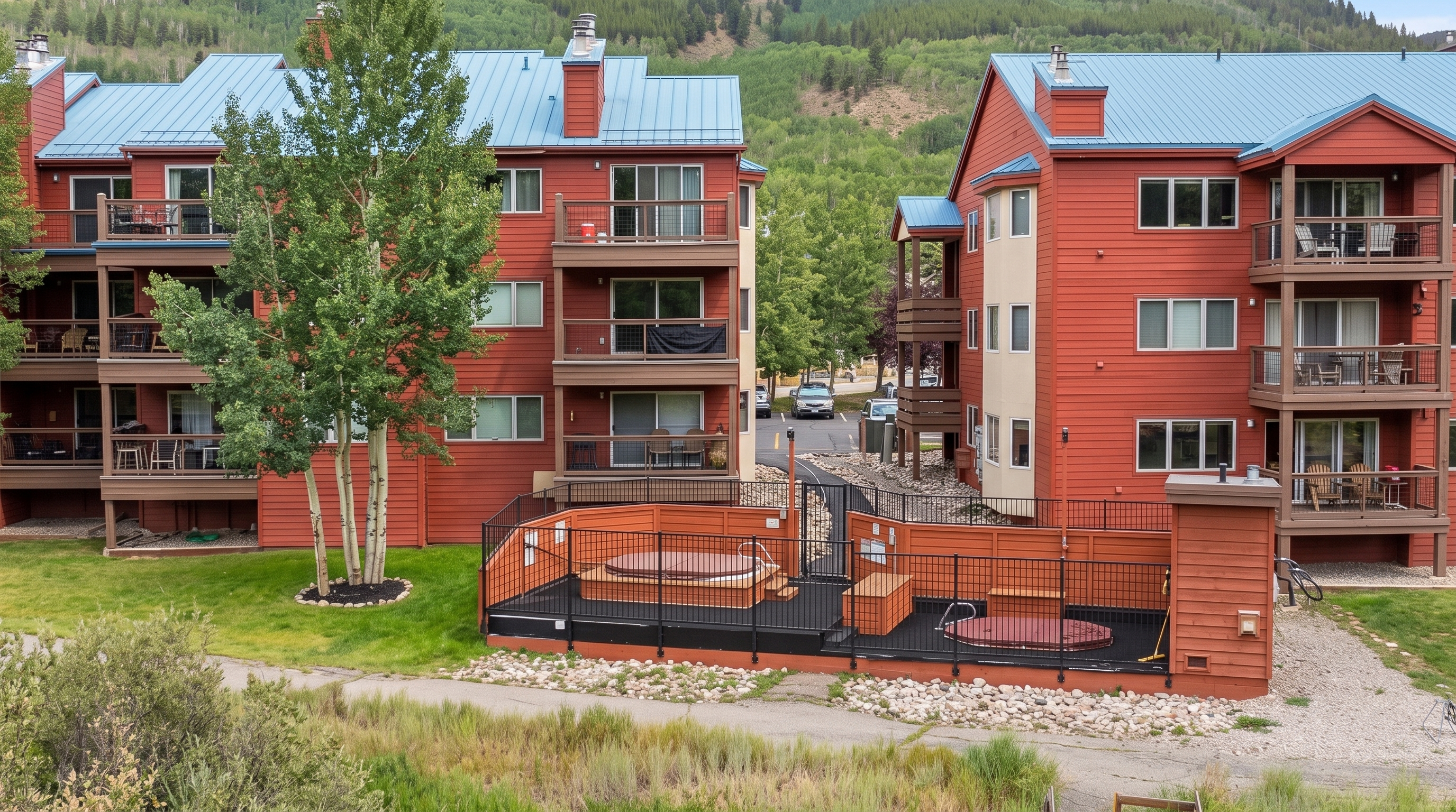 This outdoor common area features two hot tubs enclosed within a black metal fence, situated between two multi-story red buildings with blue metal roofs. The facility is set against a scenic backdrop of tree-covered mountains, providing a peaceful and recreational space for residents. The well-maintained grounds and proximity to the condominium units enhance the overall appeal of this mountain-style living community.