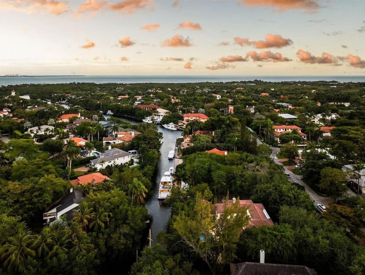 This high-angle aerial view captures a lush, tropical residential neighborhood bisected by a serene canal filled with private yachts. The landscape is densely populated with mature trees and greenery, surrounding various Mediterranean-style homes with terracotta roofs. In the distance, the horizon meets the open water under a soft, sunset-lit sky, creating a tranquil and prestigious coastal atmosphere.