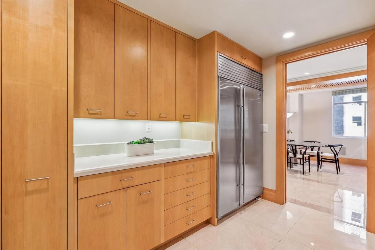 This kitchen area features light wood cabinetry with integrated appliances, including a large stainless steel refrigerator. A clean, light-colored countertop provides workspace, while a doorway leads into an adjacent dining area with a table and chairs. The space is bright and well-lit, showcasing a polished stone floor that reflects the ambient light.