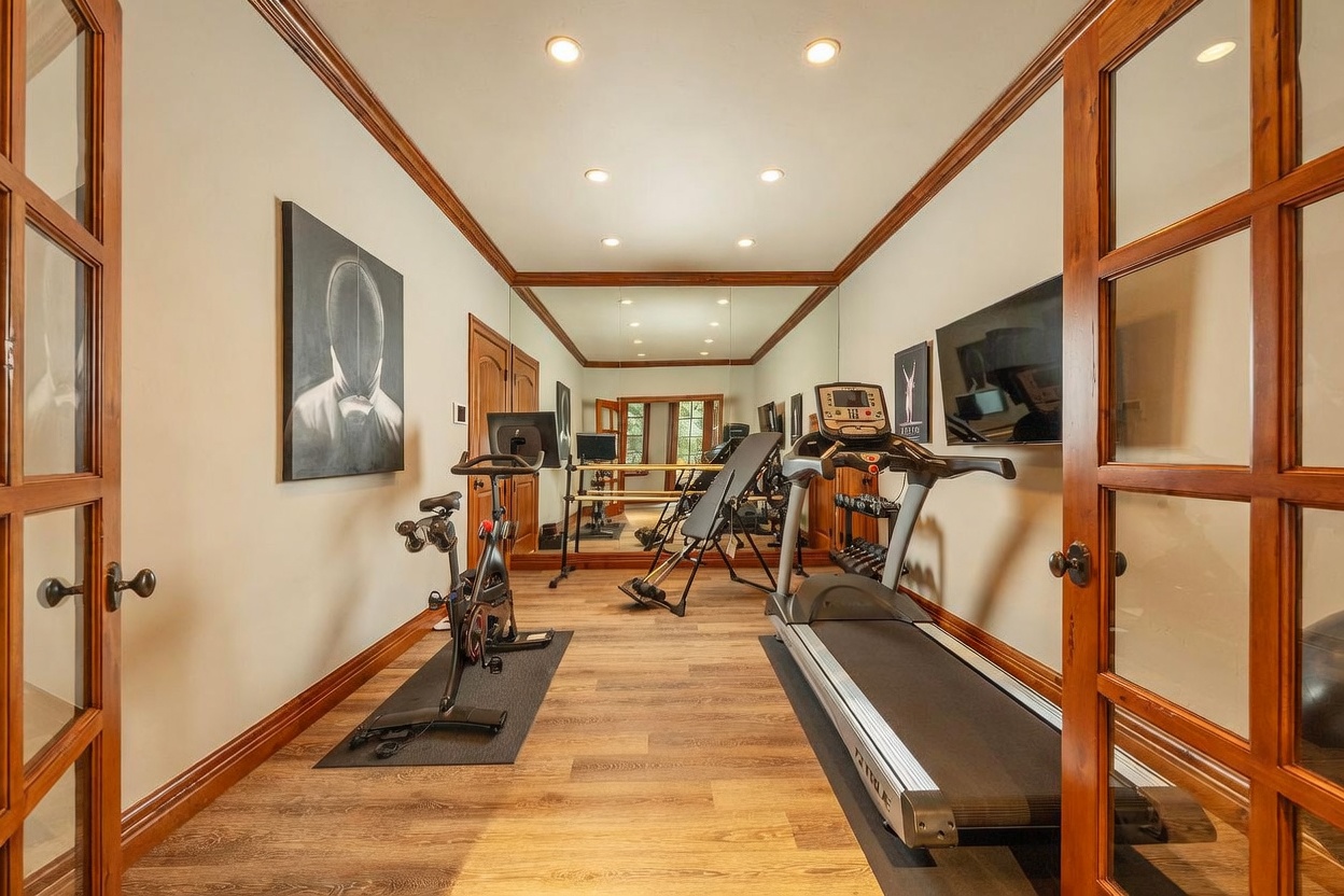 This home gym features wood-look flooring and is framed by double glass-paneled doors on both ends, creating a bright and open feel. The room is equipped with a treadmill, a stationary bike, and a weight bench, with a large wall-mounted television and a mirror that enhances the sense of space. Recessed lighting illuminates the area, which is decorated with framed artwork, providing a functional and well-lit workout environment.