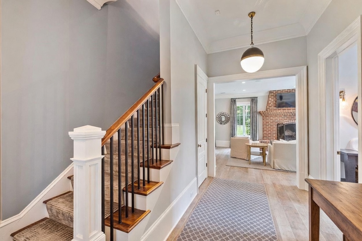 This interior view captures a bright entryway featuring a staircase with dark wood treads and black metal balusters. A patterned runner leads the eye toward an open doorway that reveals a living area with a brick fireplace and comfortable seating. The space is illuminated by a modern pendant light, creating a welcoming and airy atmosphere.