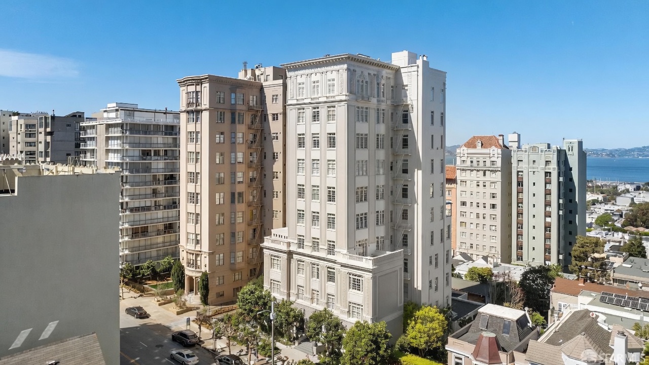 This high-angle aerial view captures a dense urban neighborhood featuring several historic, multi-story apartment buildings with classic architectural detailing. The scene highlights the interplay between the light-colored masonry facades, fire escapes, and the surrounding streetscape with mature trees and parked vehicles. In the background, a glimpse of the bay and distant hills provides a scenic context for this established residential area.