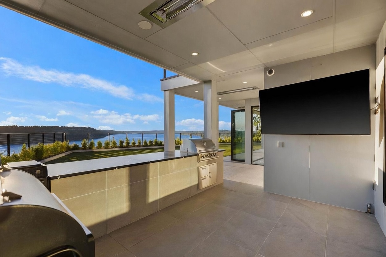This covered outdoor kitchen and lounge area features a sleek, modern design with a built-in grill and expansive counter space. A large television is mounted on the wall, creating an ideal space for outdoor entertainment, while the open side provides a panoramic view of the water and horizon. The space is finished with neutral-toned tile flooring and recessed lighting, offering a sophisticated and seamless transition between indoor and outdoor living.