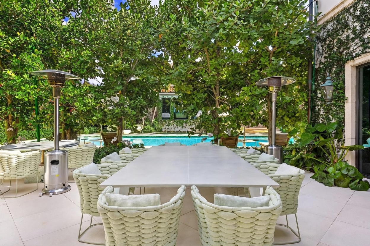 This elegant outdoor dining area features a long, light-toned table surrounded by woven chairs, set on a clean-lined stone patio. Two tall, metallic patio heaters stand on either side, while lush green trees provide a natural canopy over the space. In the background, a sparkling blue swimming pool is visible, creating a serene and luxurious atmosphere perfect for alfresco entertaining.