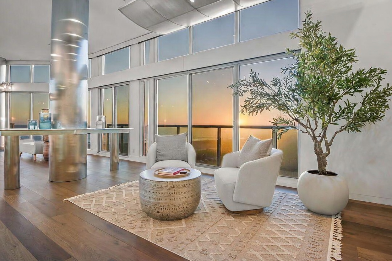 This bright, modern living area features a pair of white armchairs arranged around a metallic drum coffee table on a patterned area rug. The space is defined by expansive floor-to-ceiling windows that offer a panoramic sunset view, complemented by a sleek, polished metal structural column. A large potted olive tree adds a touch of organic texture to the minimalist, high-end interior design.