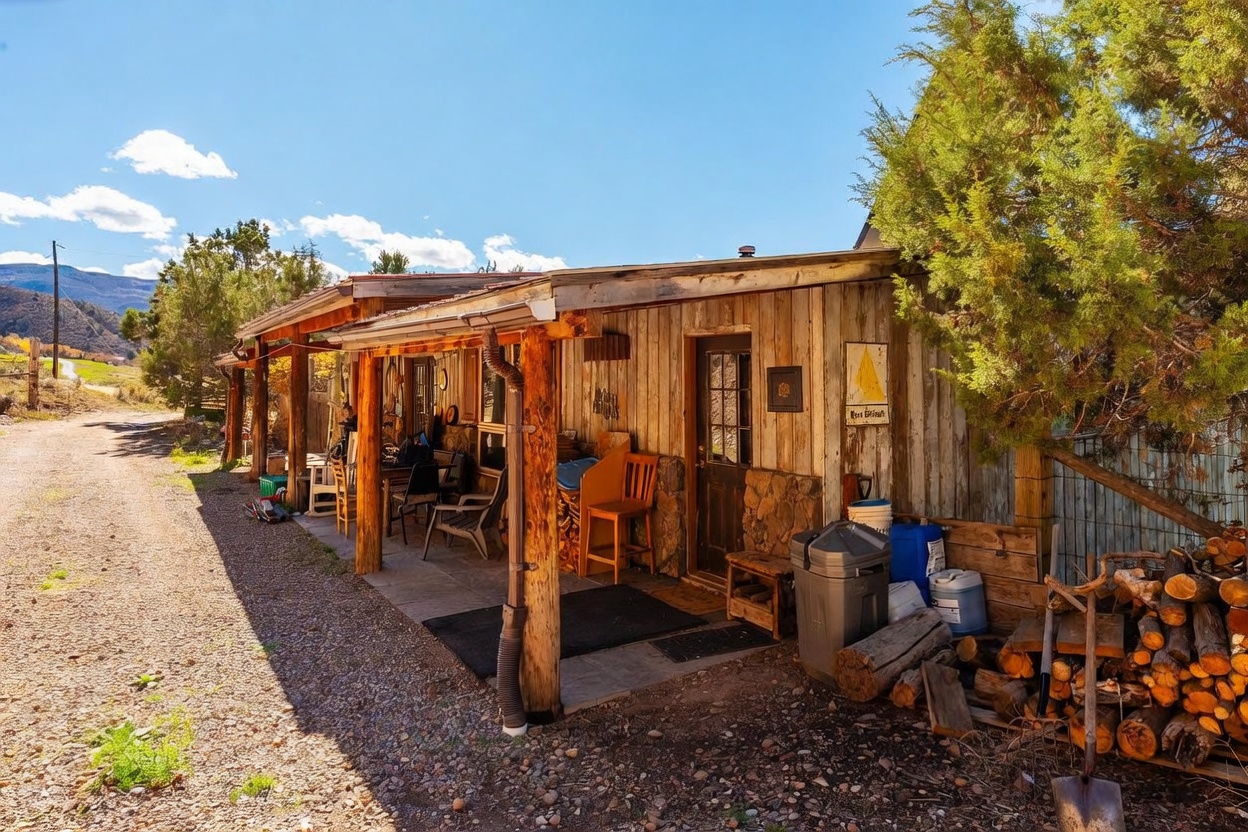 This rustic, covered porch features weathered wood siding and sturdy log support posts, creating a charming, cabin-like aesthetic. The space is furnished with simple outdoor seating and includes a stack of firewood, a trash bin, and a small sign on the wall. The perspective captures the length of the porch alongside a gravel path, with a large evergreen tree providing shade and a scenic mountain landscape visible in the distance.